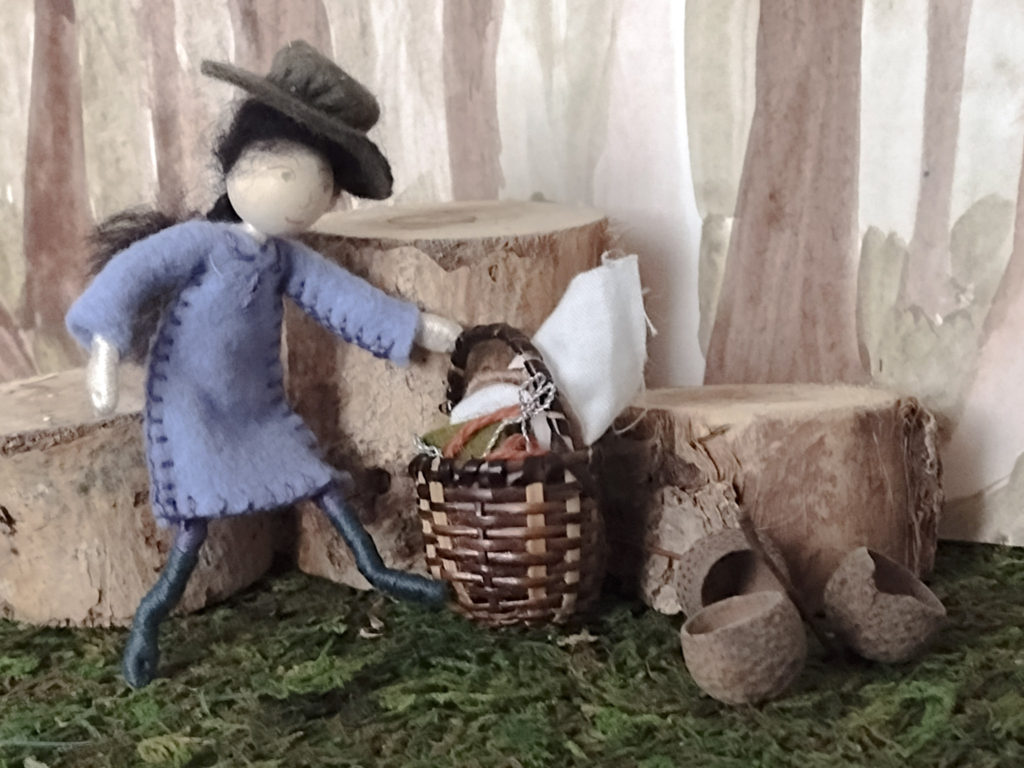 Tiny wood nymph doll, wearing a purple dress and a green sun hat bending over to pick up her embroidery basket from the green wooded ground.