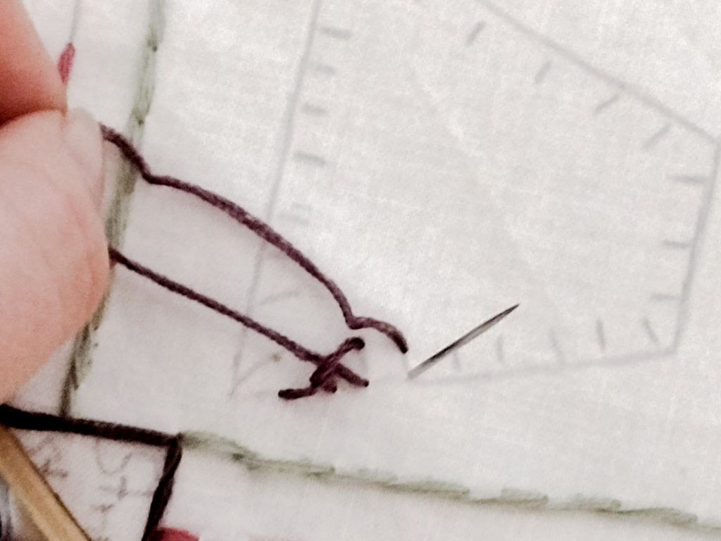 pulling embroidery needle from underneath thumb in left corner holding a loop of purple embroidery floss