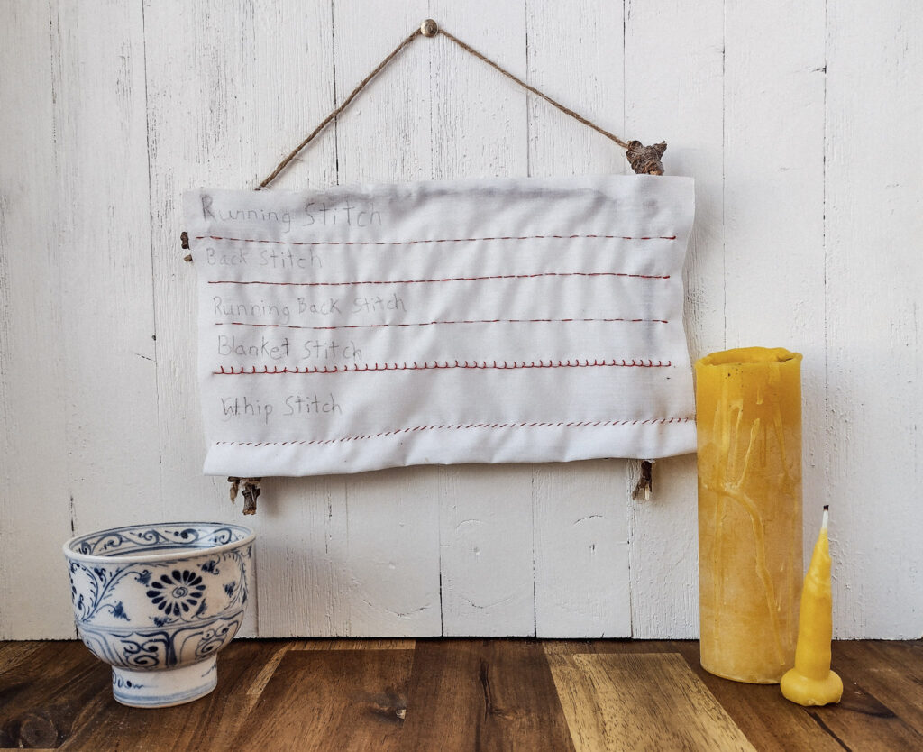 A hand sewing sampler hanging on a ship lap wall with bees wax candles and a decorative bowl